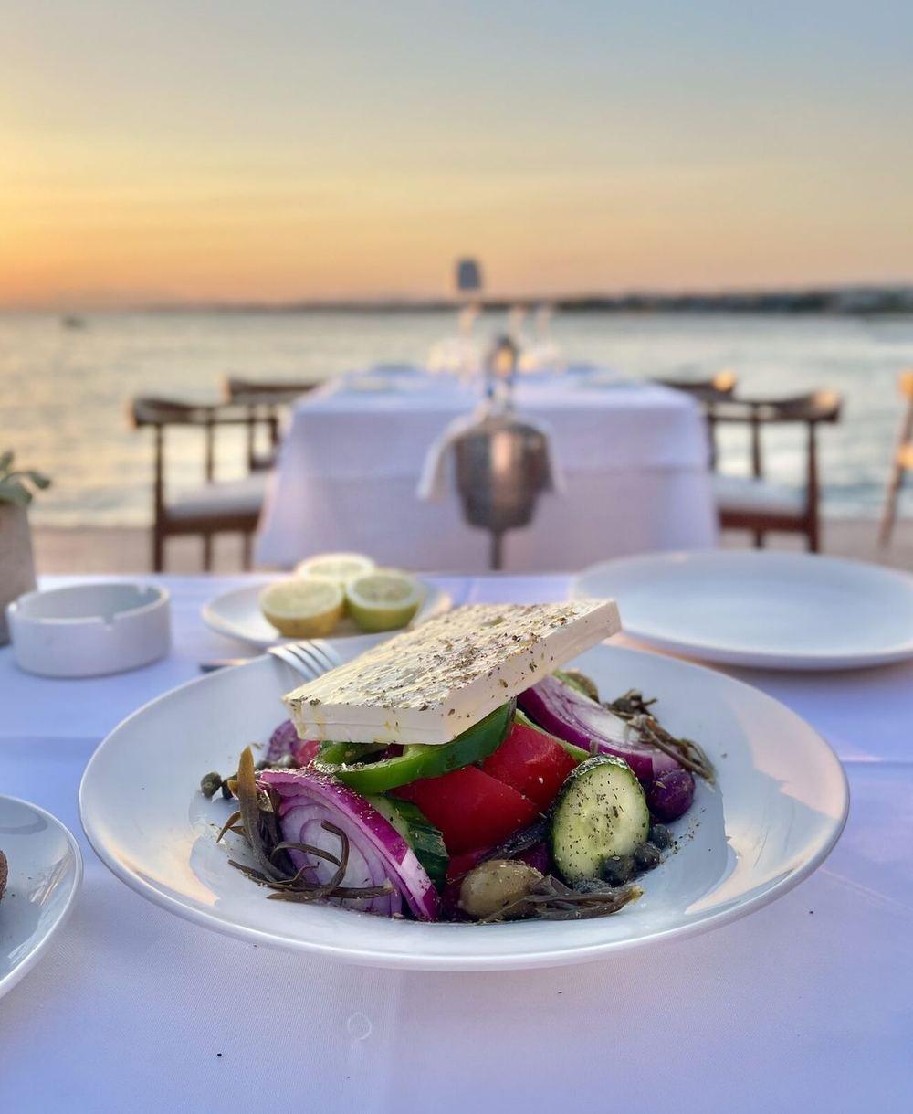 A horiatiki, the Greek village salad, on a white plate at Papaioannou Vouliagmeni, the sea and a pale sunset behind it