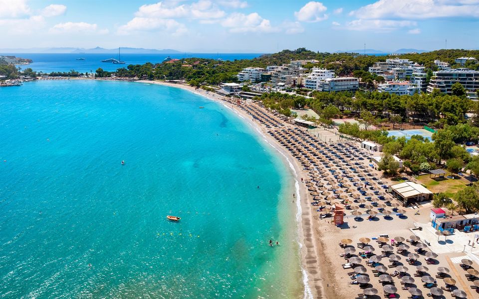 A curved bay on the coast south of Athens: turquoise shallow water, a long crescent of sand with rows of thatched umbrellas and loungers, pines and apartment blocks behind