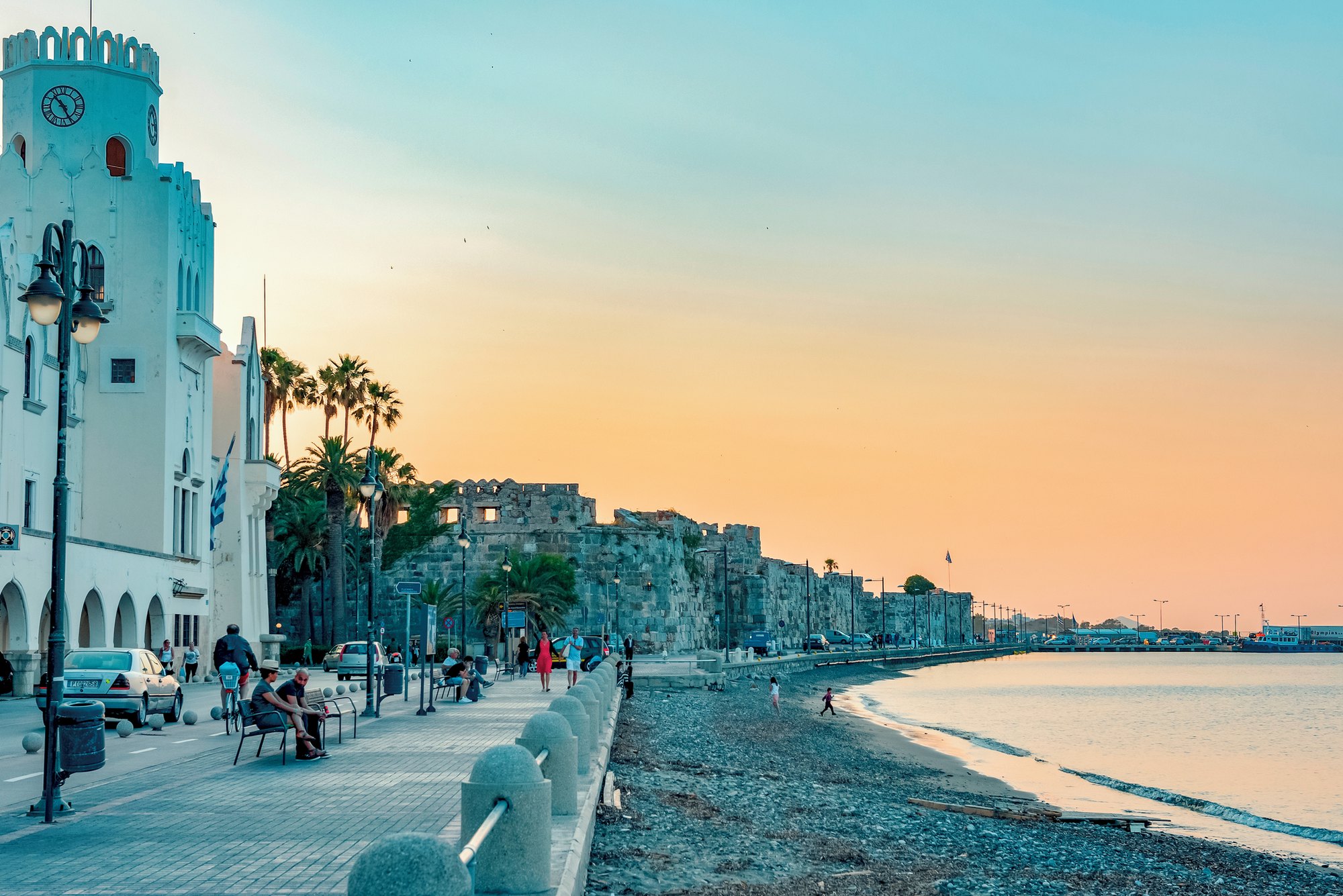 The waterfront of Kos town at dusk: palms along the shore, the Italian-built governor's palace lit, and the sea in front