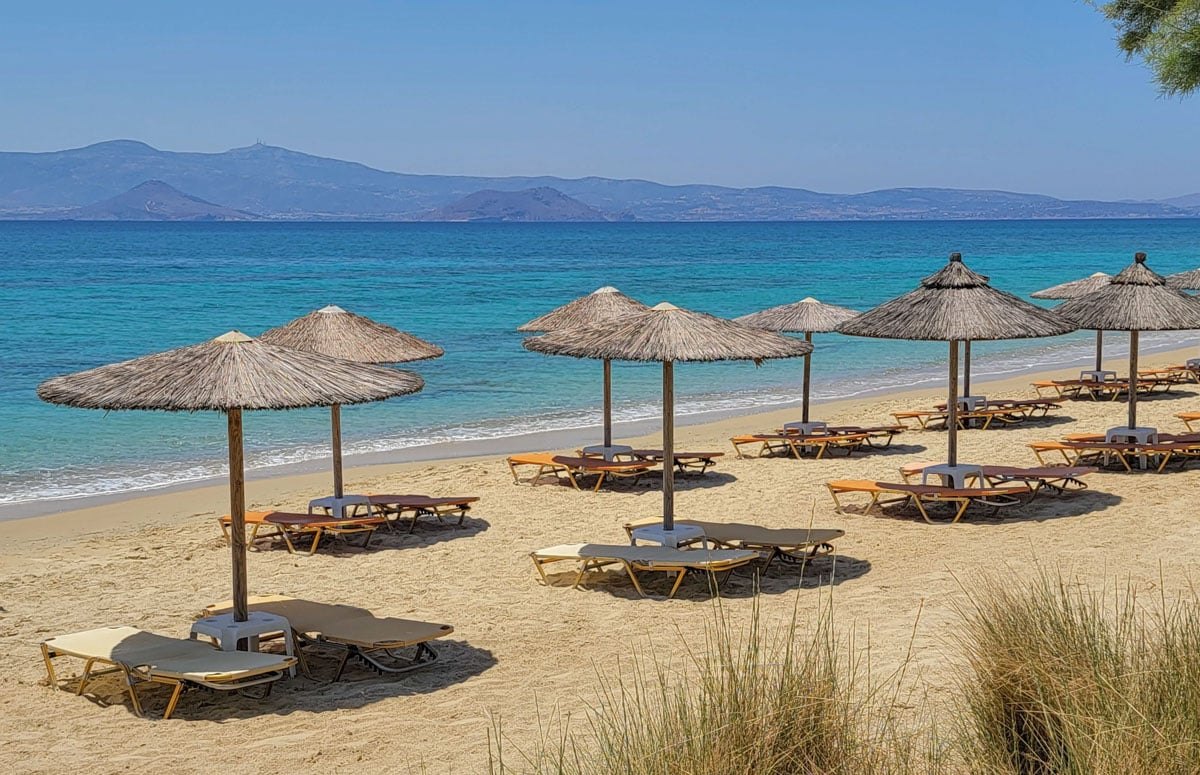 Plaka beach on Naxos: thatched umbrellas and sunbeds along a wide strip of pale sand, calm turquoise water