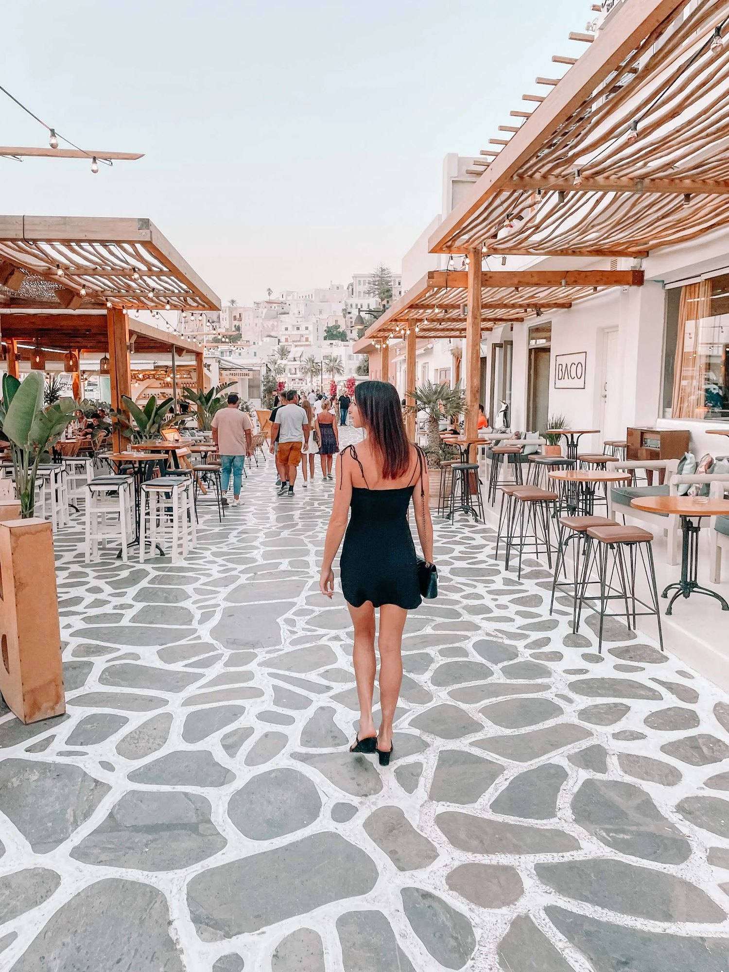 A pedestrian street in Naxos Chora at dusk with cafe tables under timber pergolas and people walking