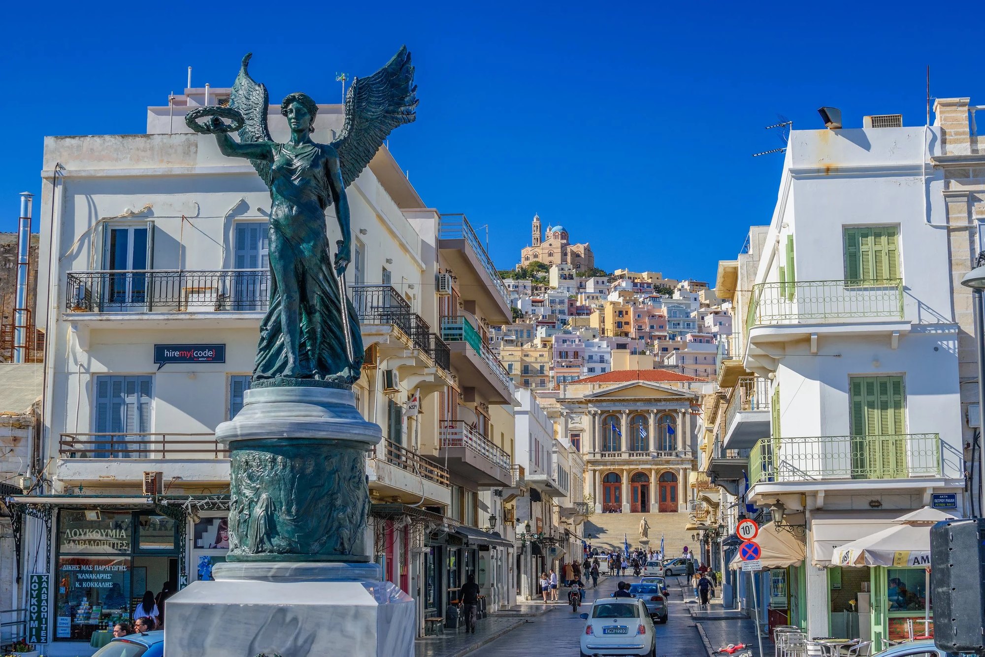 A winged bronze statue on a plinth in the middle of Ermoupoli's main shopping street, neoclassical facades either side