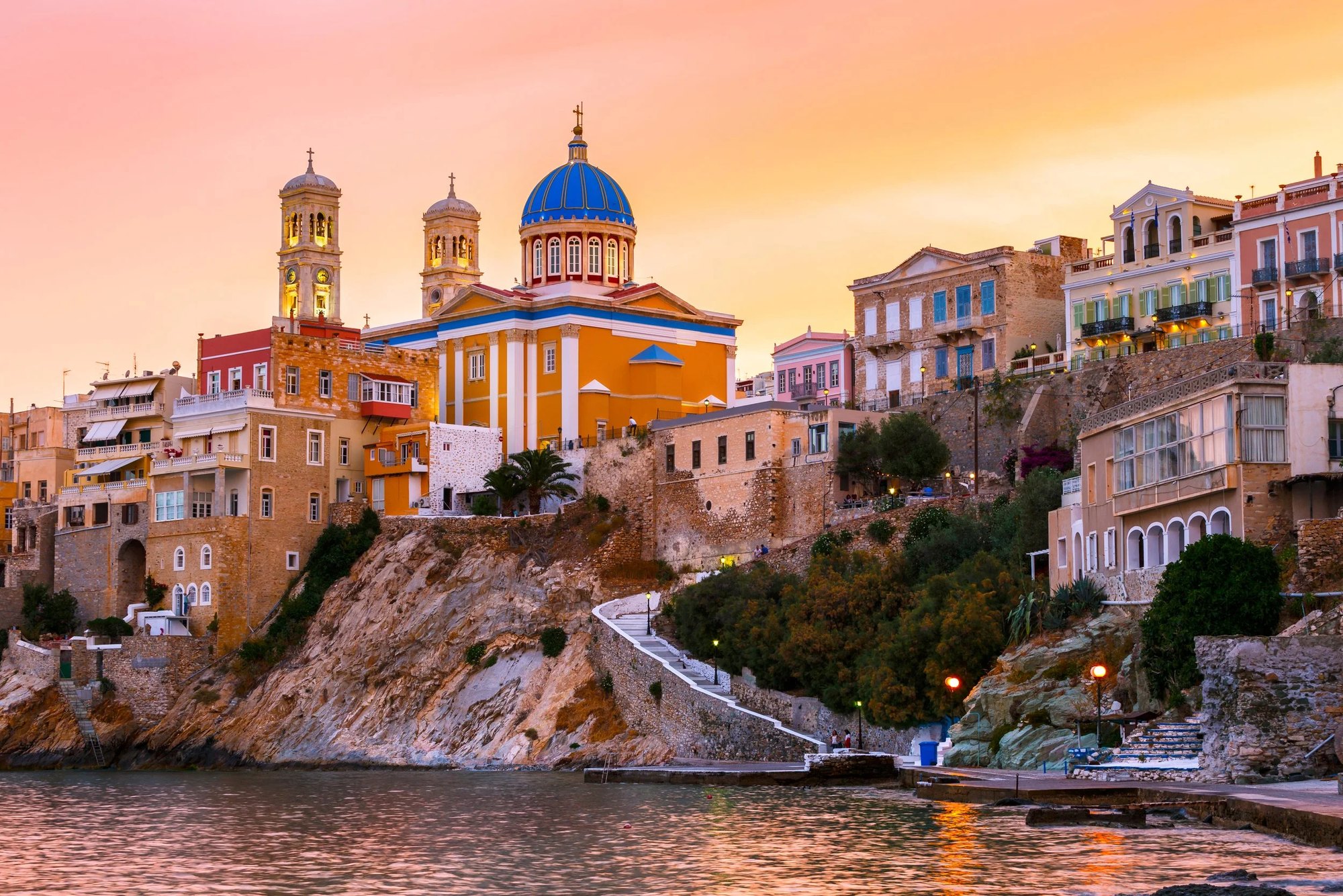 Ermoupoli at sunset seen from the water: the blue-domed church of the Dormition lit above tiers of ochre and cream mansions