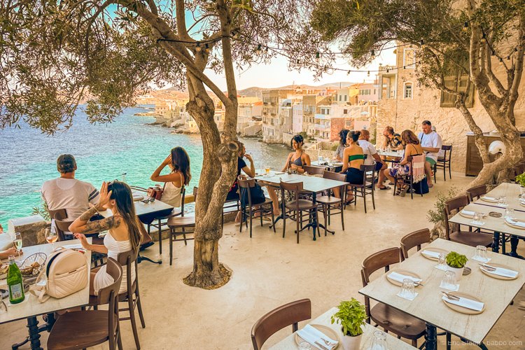 Taverna tables under a tree at the water's edge on Syros, people eating with the sea a few feet away