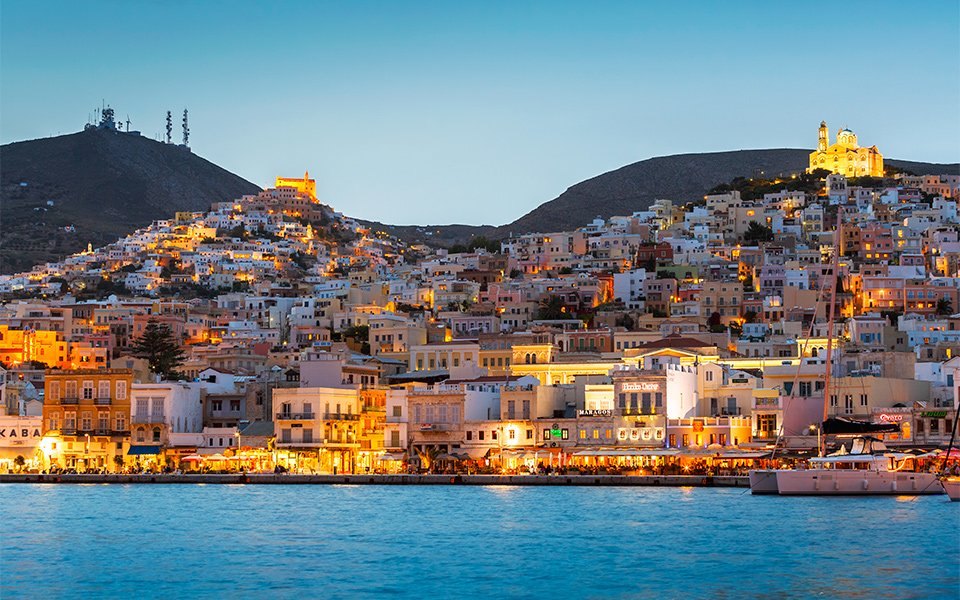 Ermoupoli from the water after dark: the lit waterfront along the harbor with two hills of houses rising behind it, a church lit on each summit