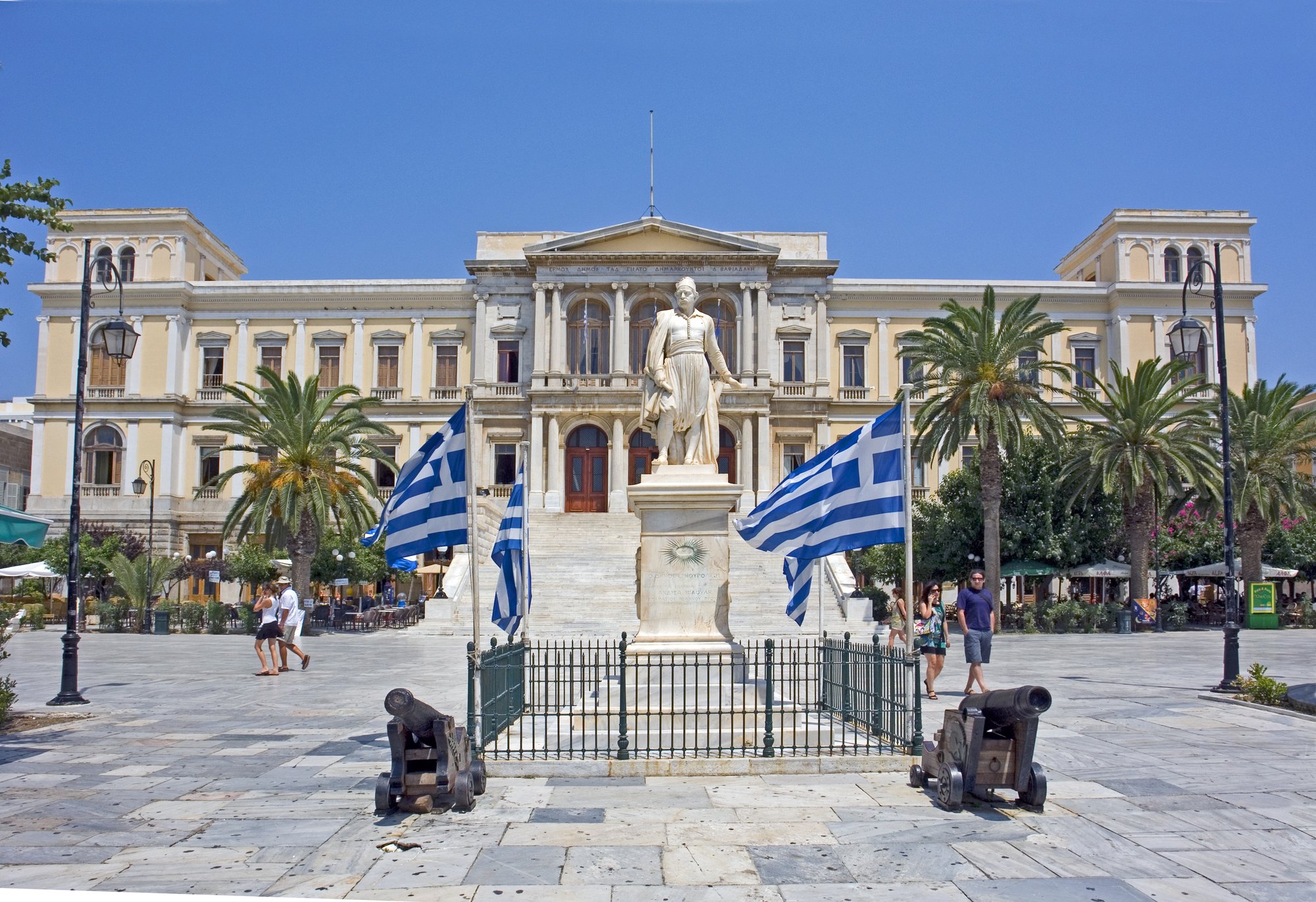 The neoclassical town hall of Ermoupoli across Miaouli Square, the statue of Admiral Miaoulis and two old cannons in front of it, palm trees and Greek flags