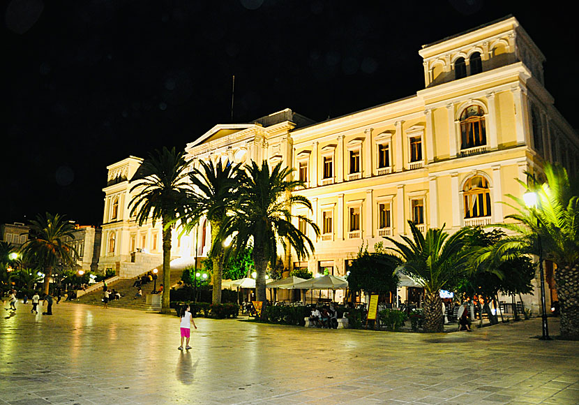 Miaouli Square in Ermoupoli at night: the neoclassical town hall floodlit behind lit palm trees, cafe umbrellas and occupied tables along one side, a child crossing the empty marble plaza in the foreground