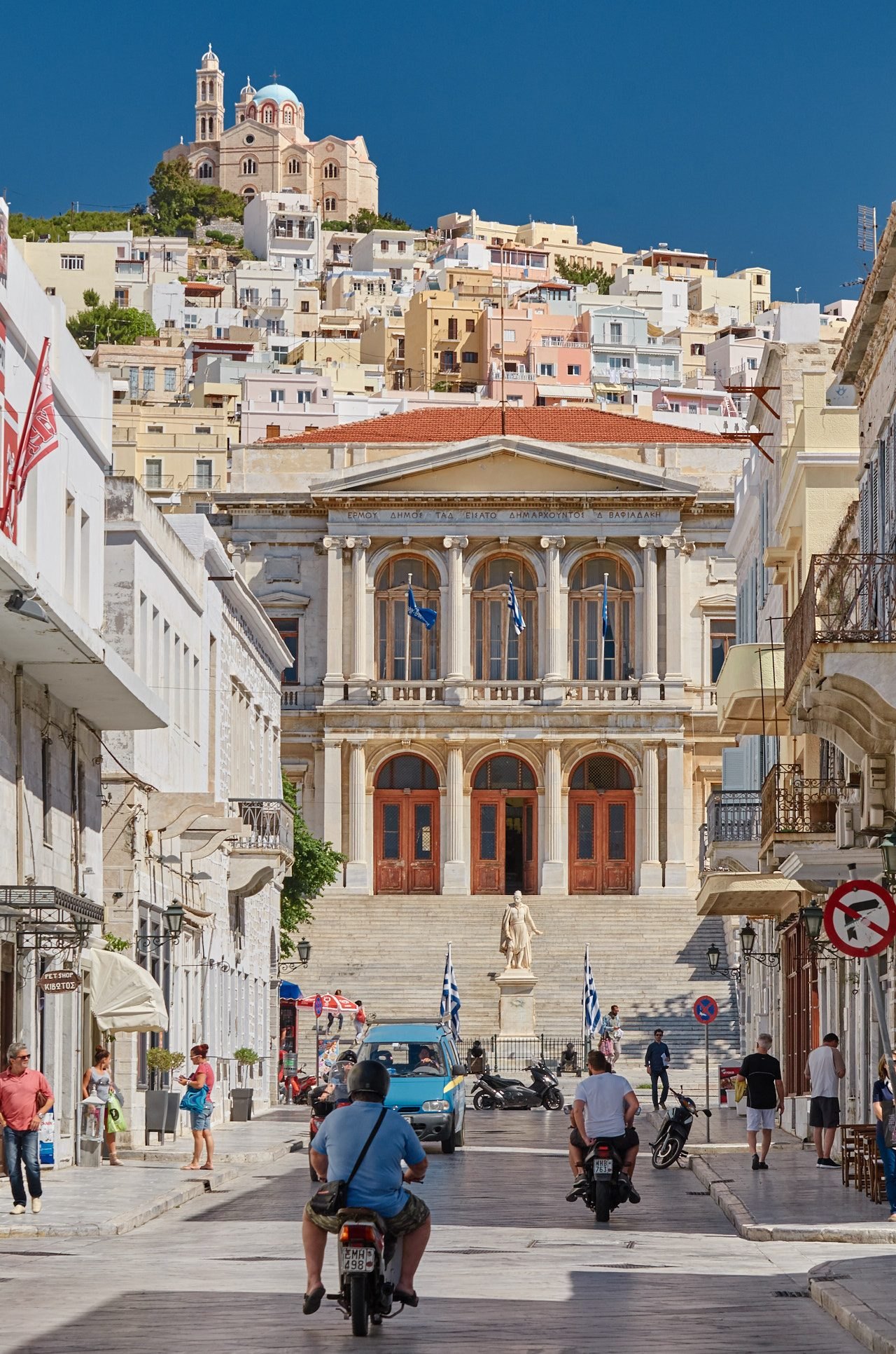 Looking up a wide marble street in Ermoupoli to the neoclassical town hall, scooters and people in the foreground, the hillside of houses behind